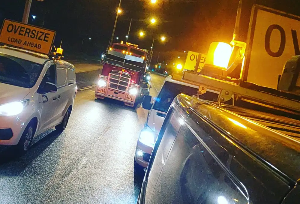 Night convoy: an oversize load approaching between two pilot vehicles under street lighting.