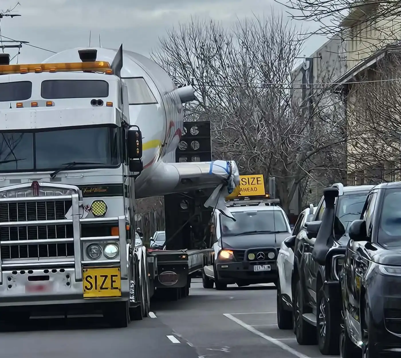 An oversize load — an aircraft fuselage on a low loader — moving down a narrow suburban street between parked cars, with a pilot vehicle alongside.